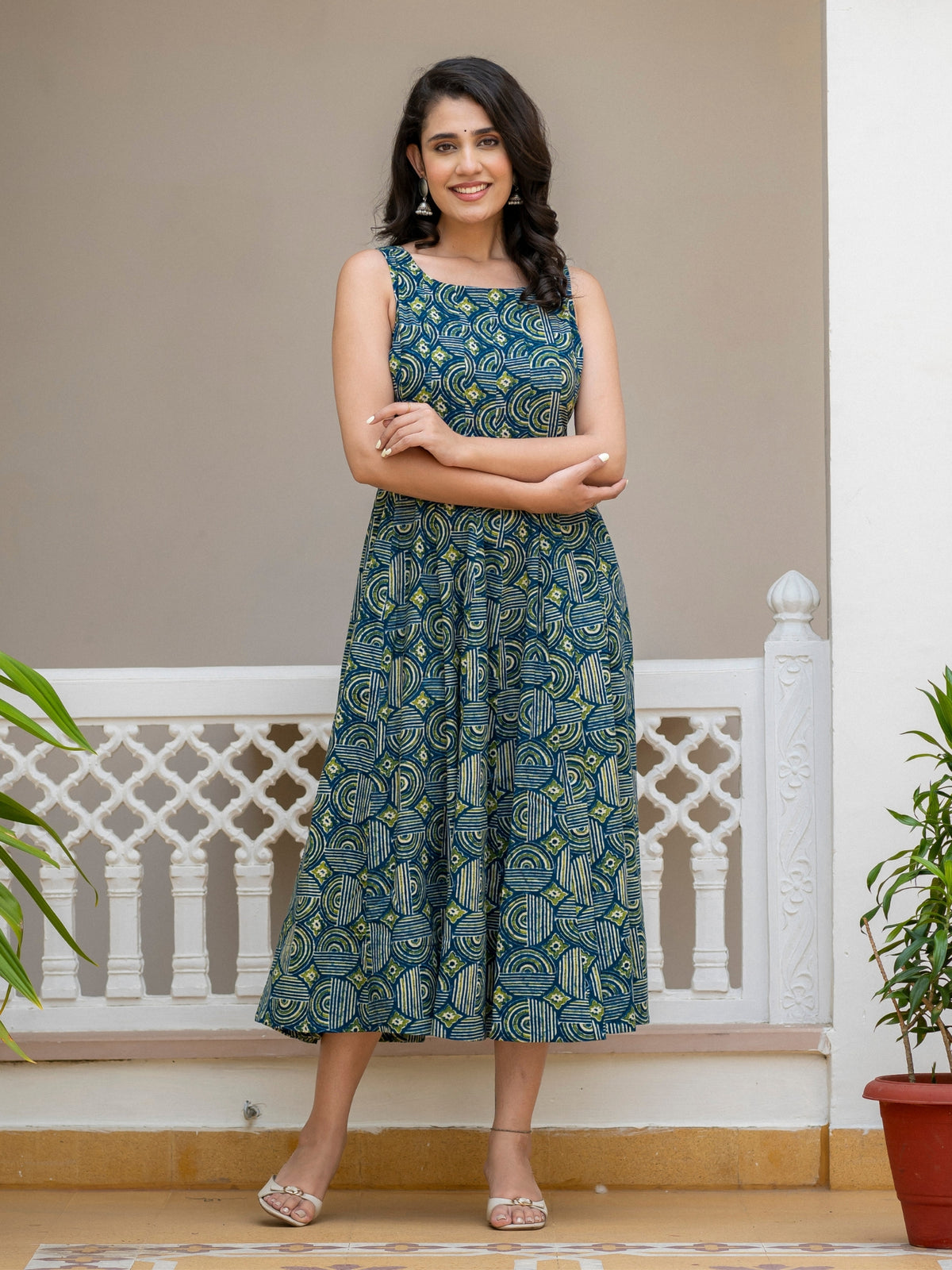 A woman modeling a sleeveless midi dress with a blue and green paisley pattern, standing in front of a white railing and beige wall.