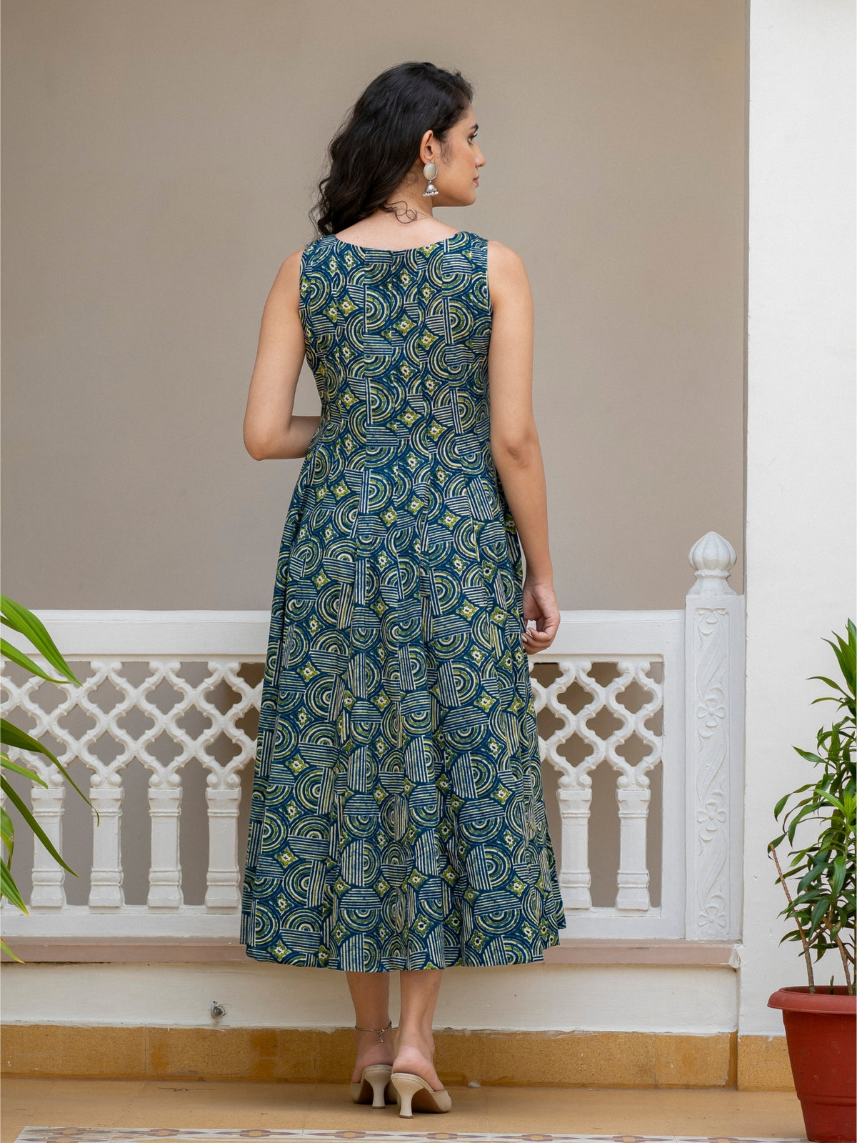 A woman stands with her back to the camera wearing a sleeveless midi dress with a blue and green paisley print, set against a beige wall and white railing.