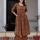 A woman stands outdoors in front of a doorway wearing a brown and rust-orange printed kurta set with matching pants. The outfit has an intricate circular and floral pattern.