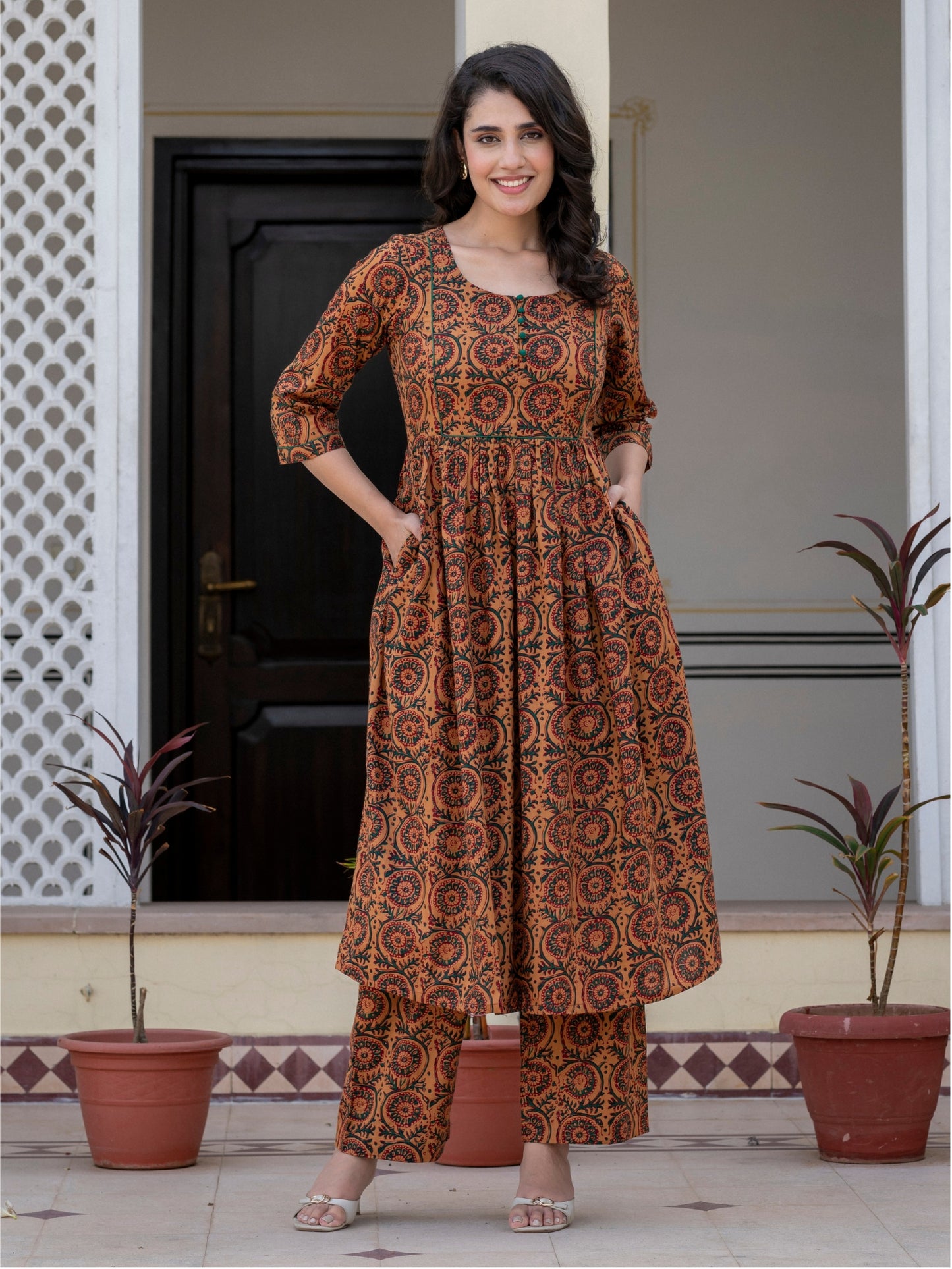 A woman stands outdoors in front of a doorway wearing a brown and rust-orange printed kurta set with matching pants. The outfit has an intricate circular and floral pattern.