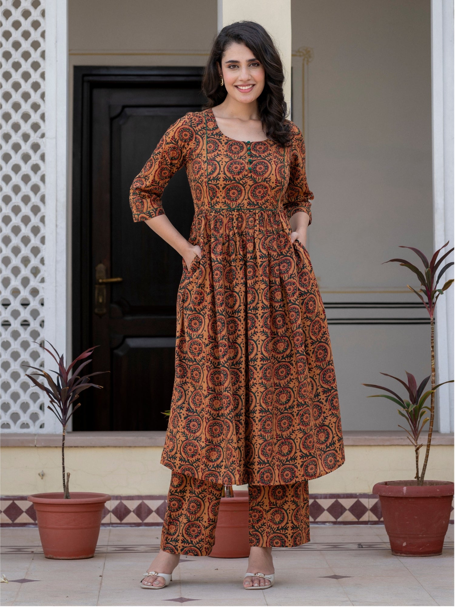 A woman stands outdoors in front of a doorway wearing a brown and rust-orange printed kurta set with matching pants. The outfit has an intricate circular and floral pattern.