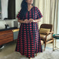 A woman wearing a black and red patterned dress stands in a living room setting. The dress has a V-neckline and short sleeves, featuring a repeating red semicircle motif on a black background.