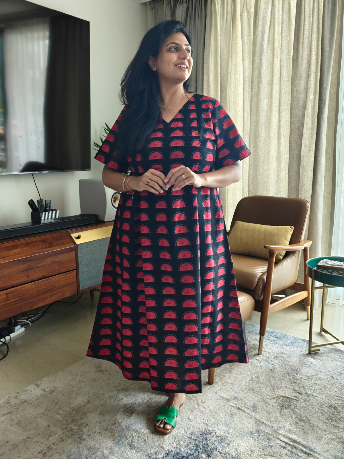A woman wearing a black and red patterned dress stands in a living room setting. The dress has a V-neckline and short sleeves, featuring a repeating red semicircle motif on a black background.
