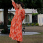A woman in a vibrant orange ikkat printed dress turns slightly, offering a side-back view in a serene garden setting.