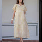 A woman models a short-sleeved midi dress with vertical mustard yellow and white stripes, standing in front of a light-colored building wall.