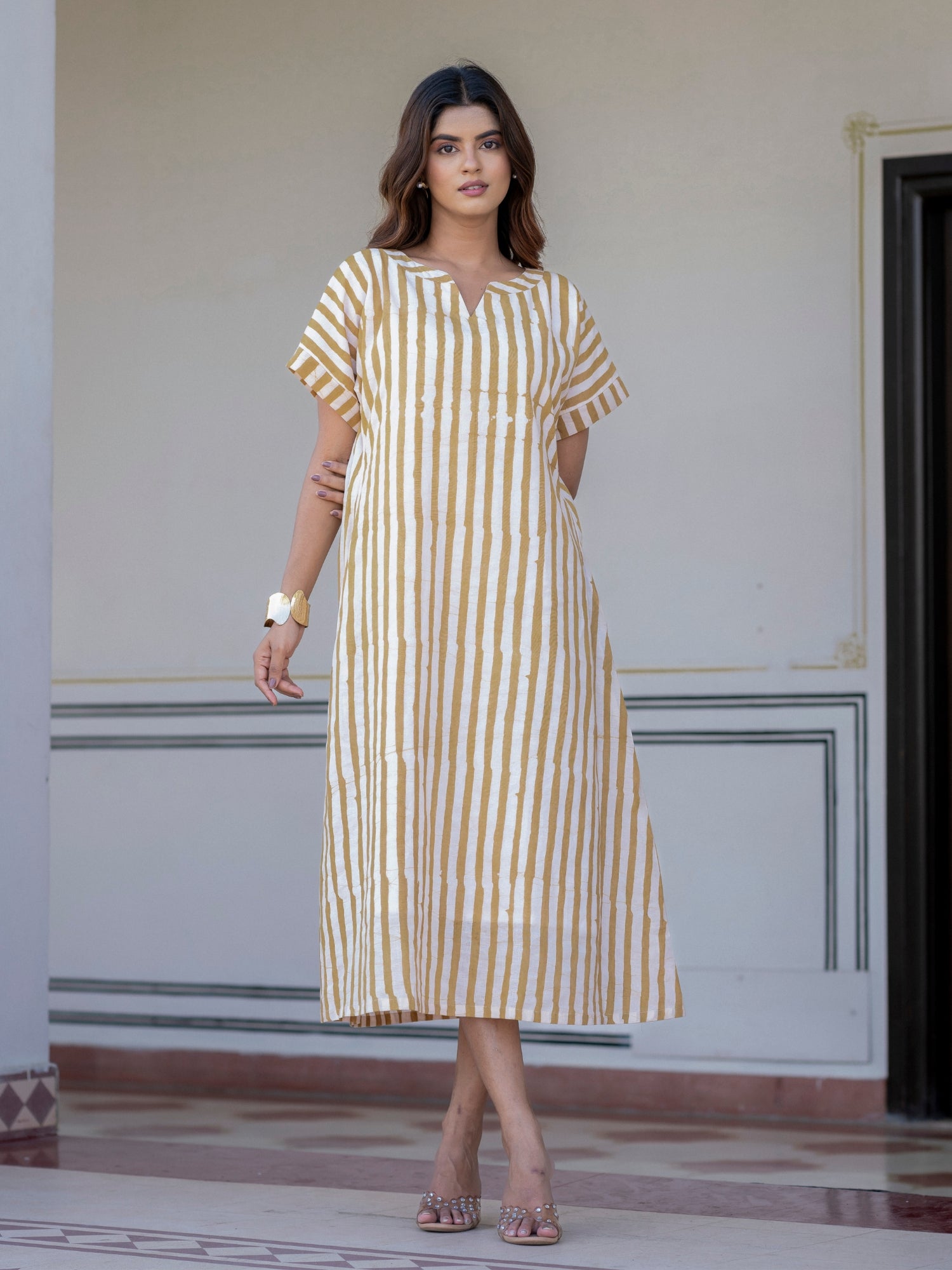 A woman models a short-sleeved midi dress with vertical mustard yellow and white stripes, standing in front of a light-colored building wall.