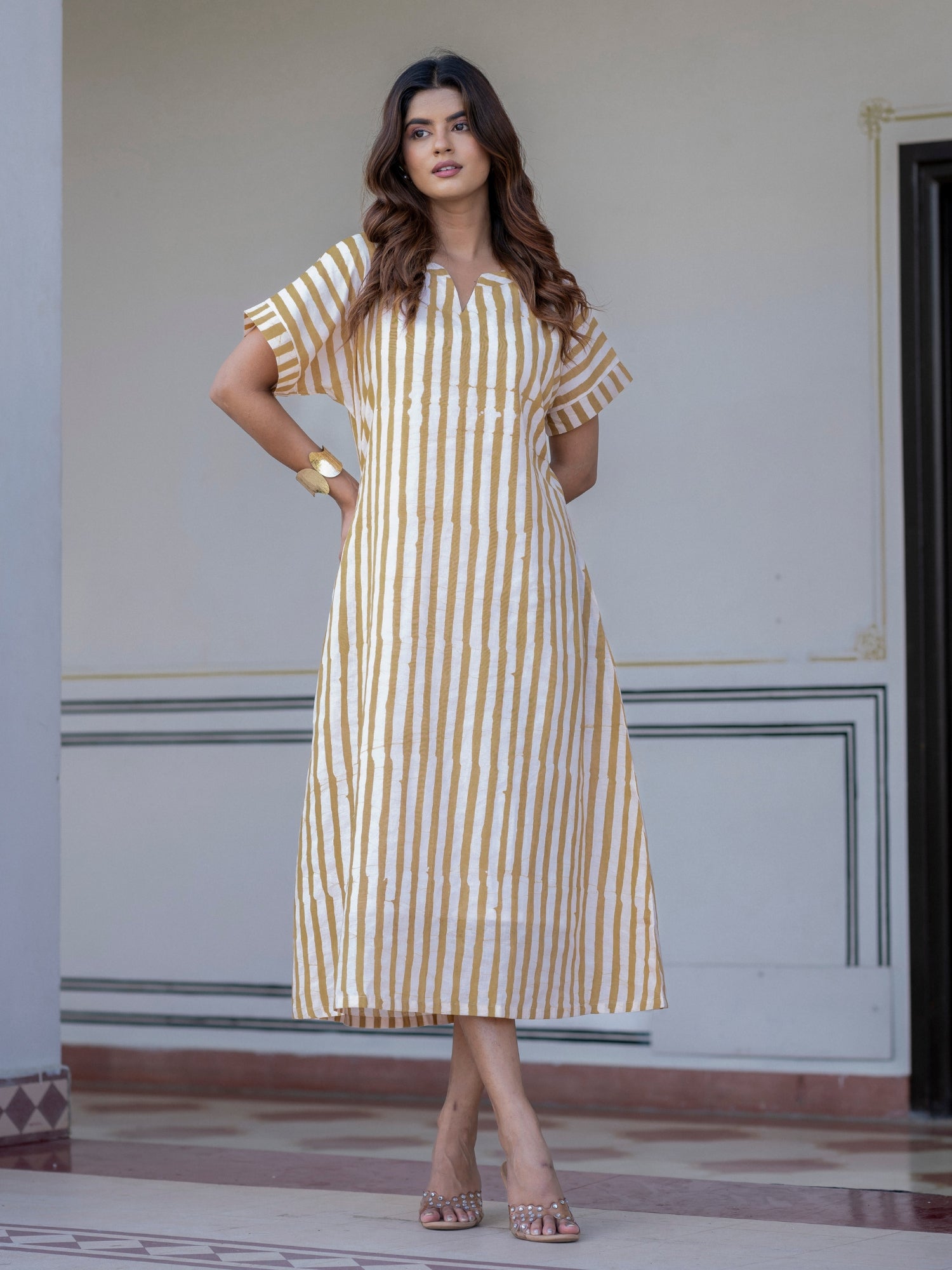 A woman modeling a short-sleeved midi dress with vertical mustard yellow and white stripes, standing in front of a light-colored wall.
