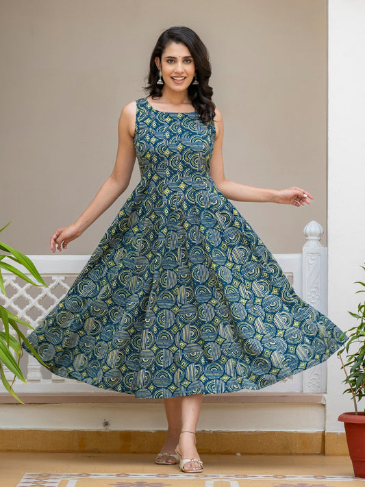 A woman modeling a sleeveless, knee-length A-line dress with a blue and green paisley pattern, standing in a doorway with her hands outstretched to show the dress's flare.