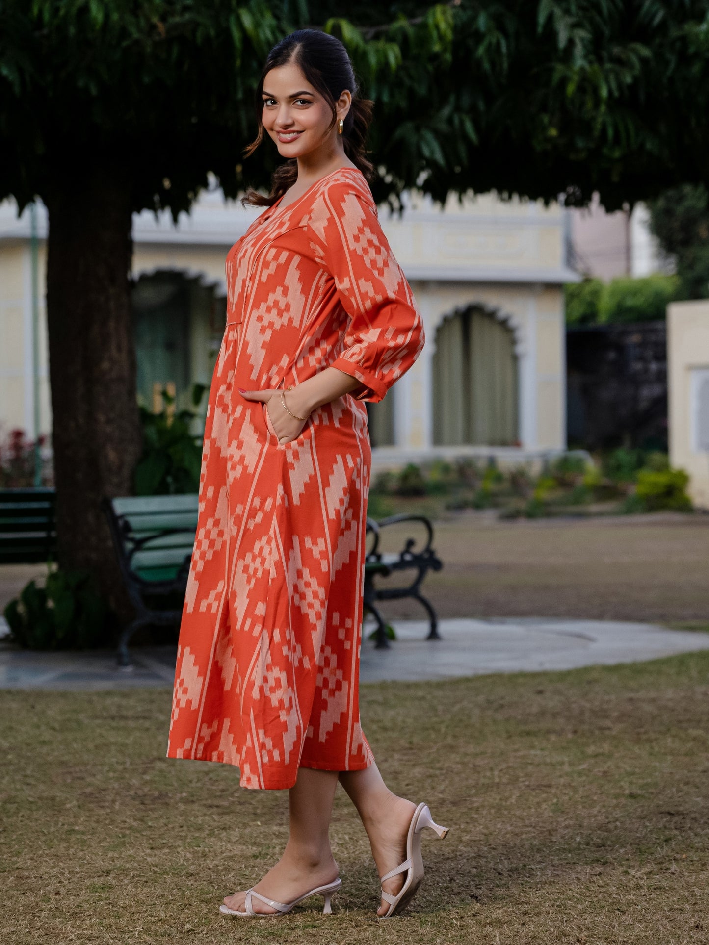 Playful pose of a woman wearing an orange ikkat dress, with the outfit’s flare and print clearly visible in an open garden area.