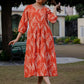 Full-length outdoor photo of a woman in an orange ikkat printed dress, standing on grass with trees and heritage-style architecture in the background.
