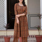 A woman stands outdoors in front of a doorway wearing a brown and orange patterned kurta with matching pants. The outfit has an intricate circular geometric print in shades of brown, orange, and black.