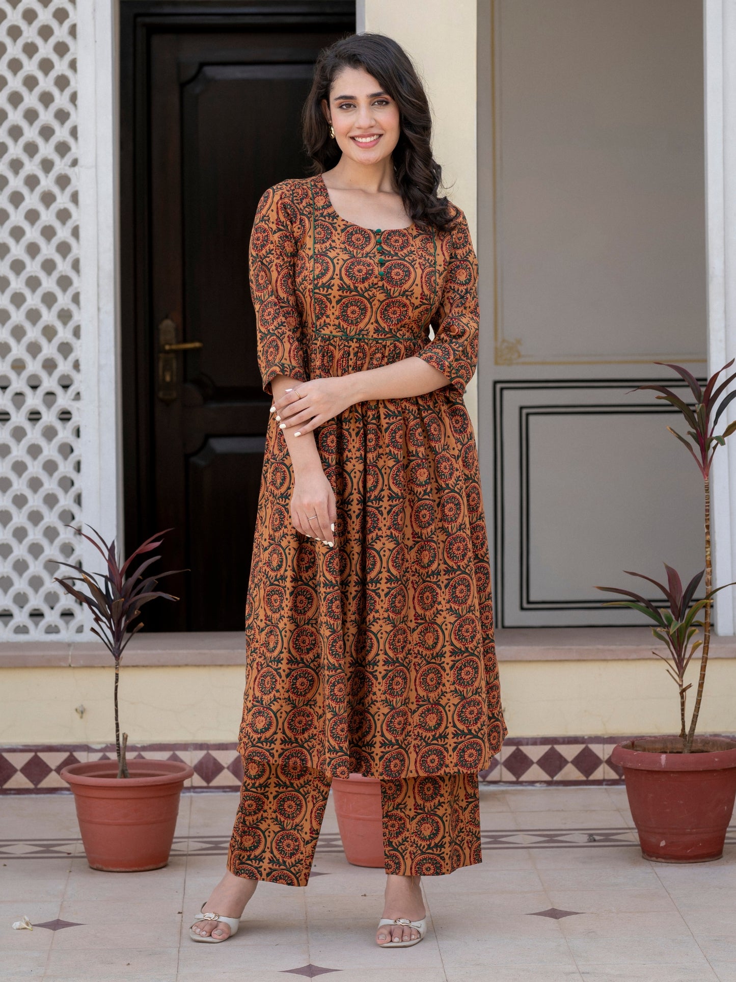 A woman stands outdoors in front of a doorway wearing a brown and orange patterned kurta with matching pants. The outfit has an intricate circular geometric print in shades of brown, orange, and black.